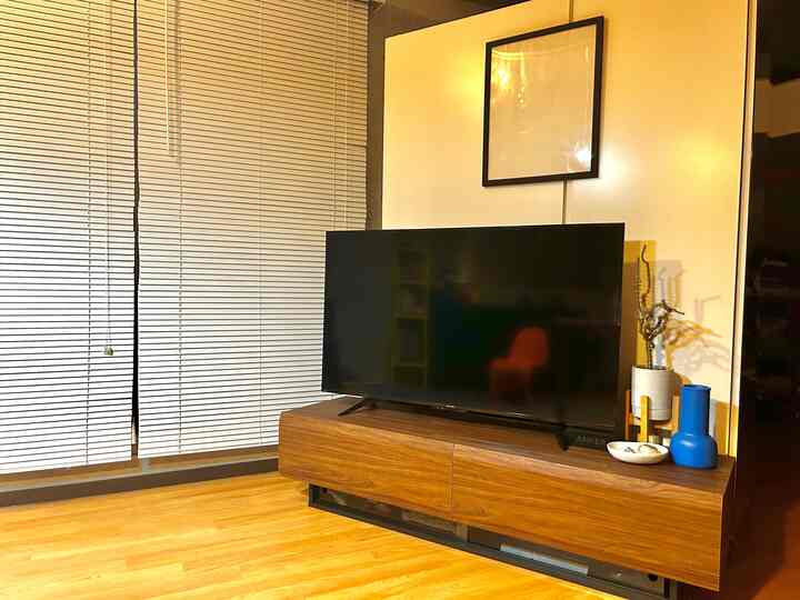 暖色の照明が印象的なワンルームのリビング、ダークブラウンのテレビ台と白いブラインドが調和したナチュラルな空間