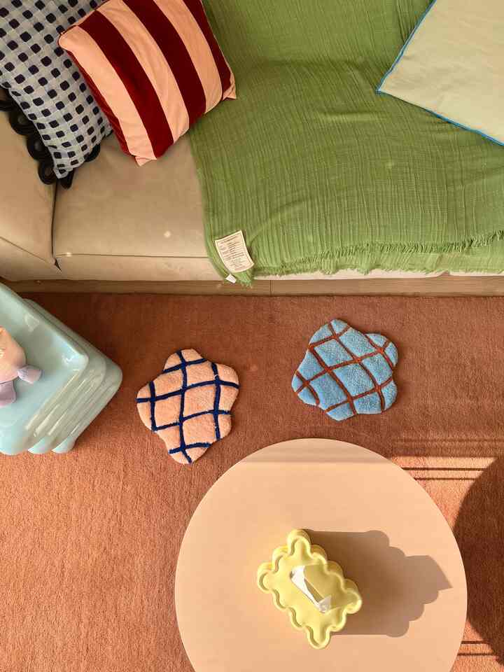 温かみのあるナチュラルトーンのリビングルーム、ベージュのソファと茶色のラグ、個性的なクッションと丸いコーヒーテーブルが配置された印象的な空間