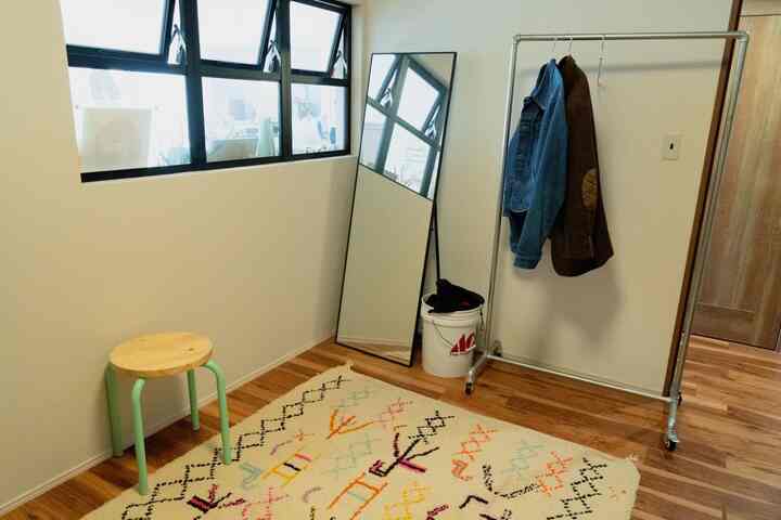 明るい白い壁と木製床の小さなウォークインクローゼット、スツールと全身鏡が配置された実用的な空間