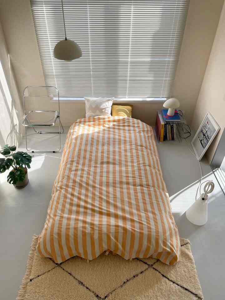 ナチュラルカラーの寝室、ストライプのベッドカバーと透明な折りたたみ椅子が配置されたミニマルで印象的な空間