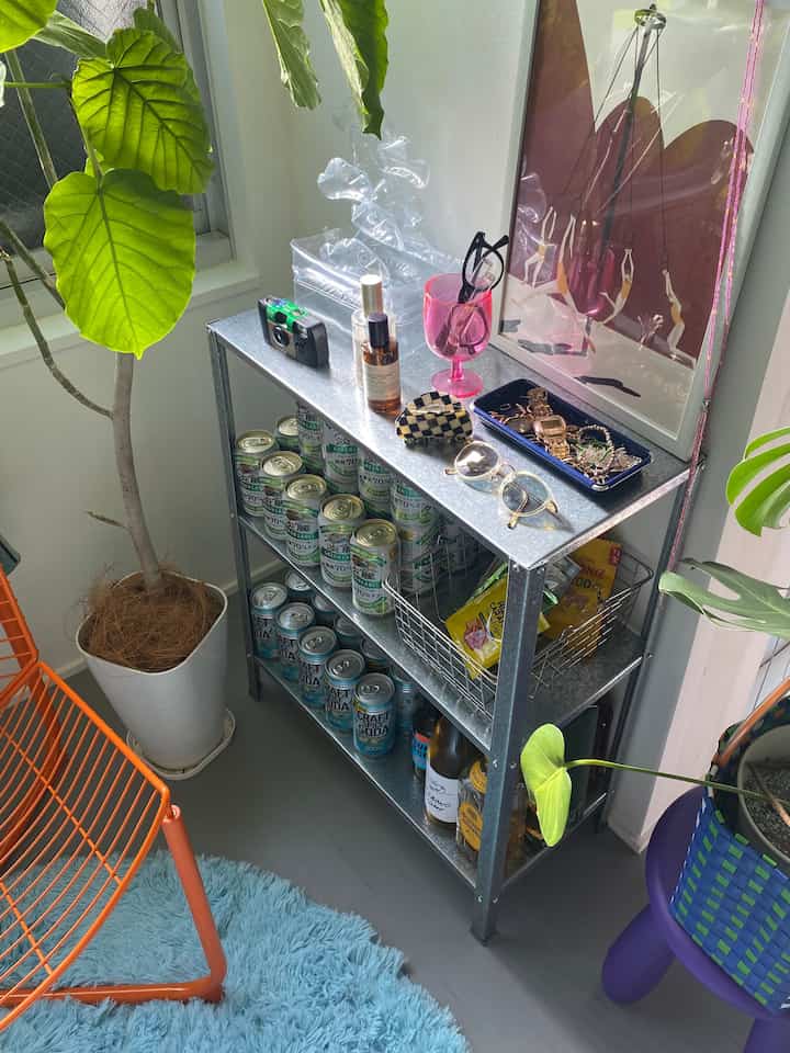 緑の植物とオレンジのワイヤーチェアが配された狭くて居心地の良い部屋、飲み物や装飾品が置かれた金属製の棚付き