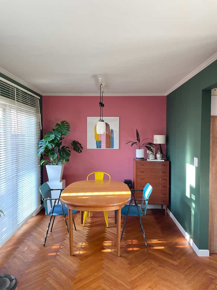 Dining room with a pink central wall flanked by green walls, featuring a wooden dining table and colorful chairs in a natural setting