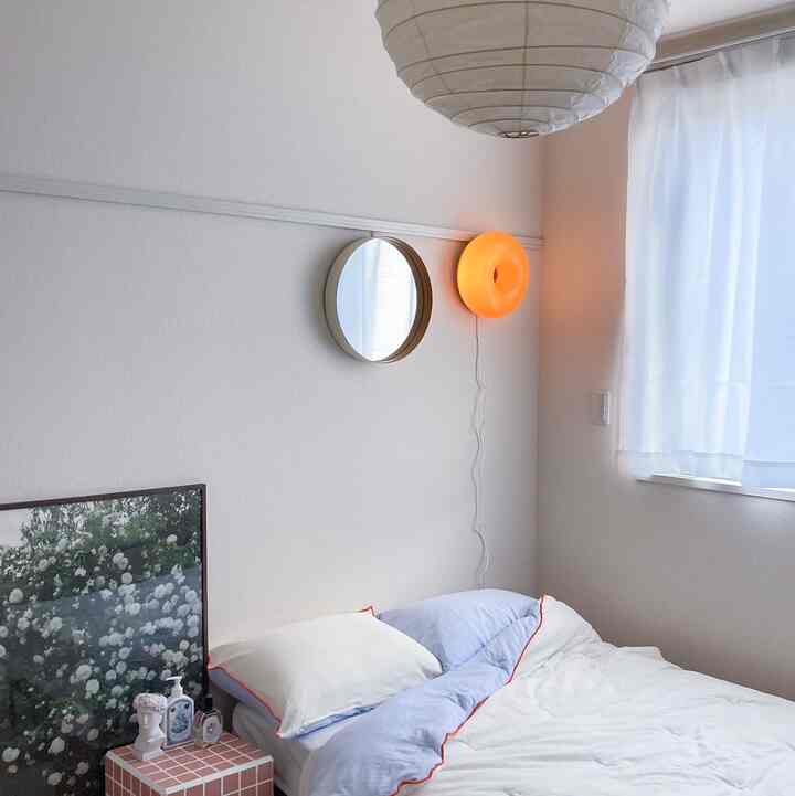 ホワイトとベージュの色調の寝室、ブルー縁取りの寝具、ピンクのタイルのナイトテーブル、丸い壁掛け鏡、暖かなオレンジ色の壁照明が配置された雰囲気のあるインテリア