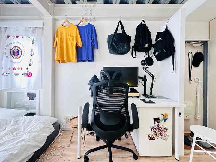 白い壁と木目調の床のある狭いワンルームのホームオフィス空間、黒いオフィスチェアとデスク、壁に掛けられた青と黄色のシャツが調和したシンプルなインテリア