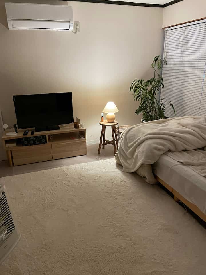 A white and brown toned bedroom featuring a wooden nightstand lamp and plant, creating a cozy modern atmosphere