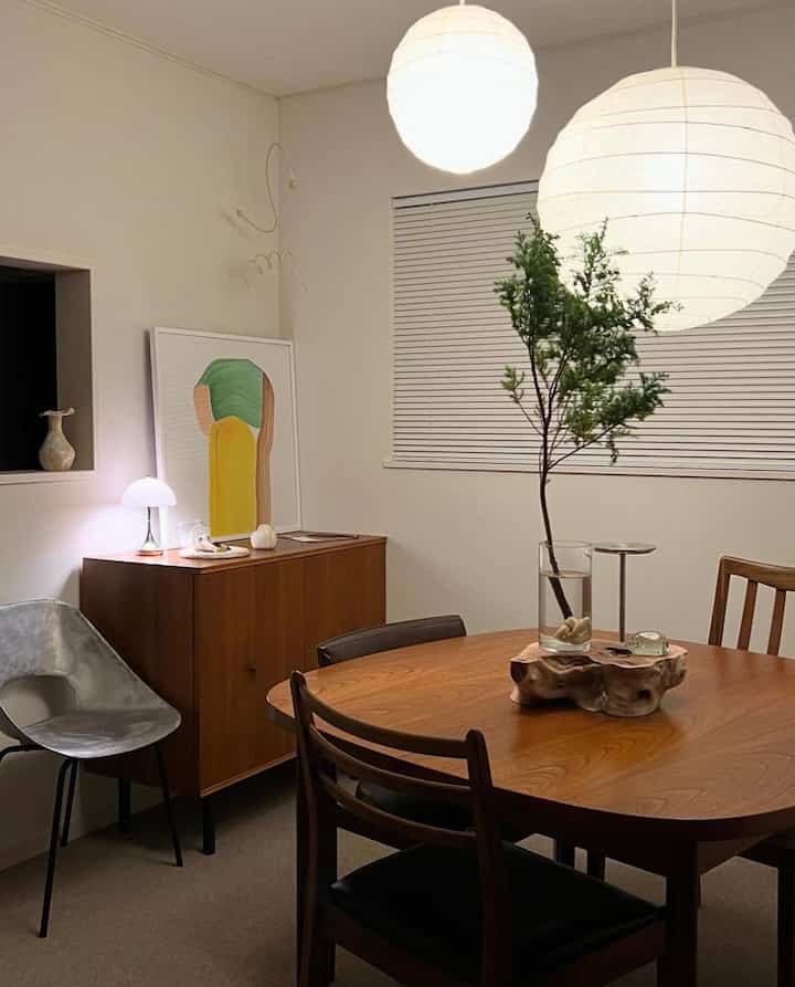 A white-toned dining room featuring a mid-century modern brown wooden dining table and chairs with warm lighting accents