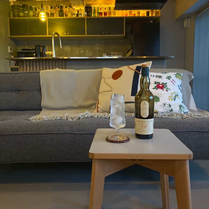 ナチュラルモダンなリビングルーム、グレーのソファと木製テーブル、ウイスキーボトルとハイボールグラスを配し、温かみのあるゴールドの照明が印象的な空間。