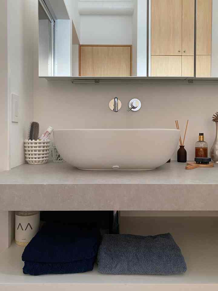 白とグレーの色調のバスルーム、中央に洗面台、青いタオルがきれいにたたまれ、ディフューザーとハンドウォッシュが配されたシンプルで落ち着いたインテリア