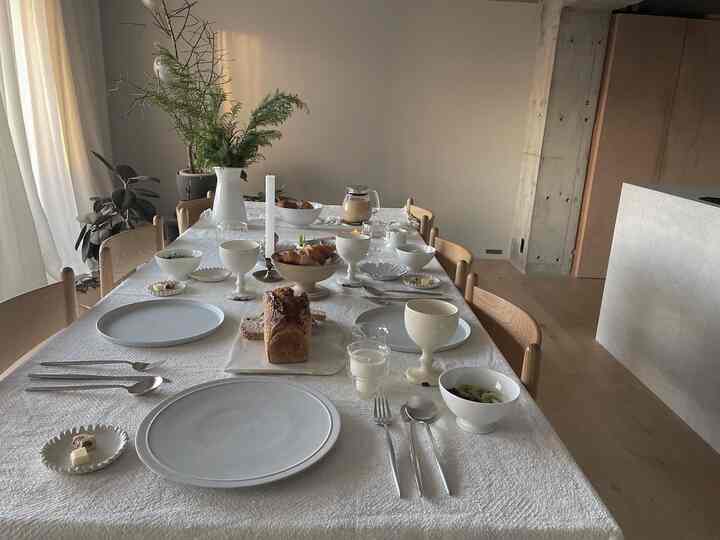 ナチュラルで明るい色調のダイニングルーム、白いテーブルクロスにカトラリーや皿、植物が配置されたシンプルな朝食の空間