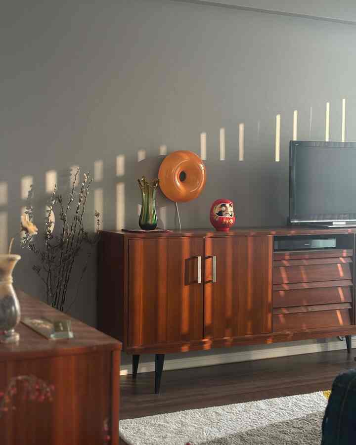 Warm wood-toned mid-century modern living room featuring an orange table lamp, a wooden cabinet, and vertical blinds casting shadows on the wall
