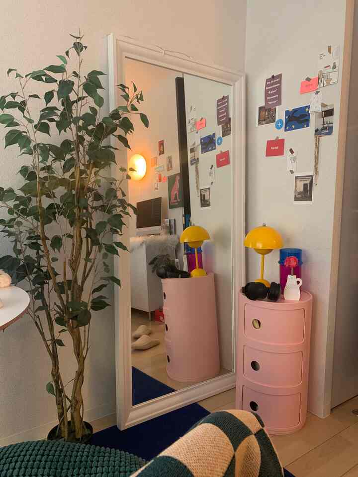 ピンクの収納と黄色のランプが置かれた6畳のワンルーム、壁の鏡と植物が配置された雰囲気のあるインテリア
