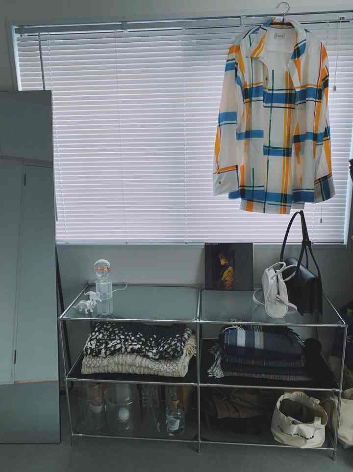 白いブラインドを背景にしたコンパクトな部屋、モジュール式の金属棚にニットやバッグが収納され、自然な雰囲気のあるインテリア