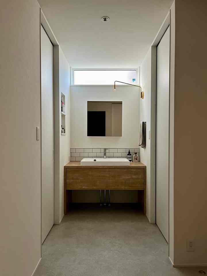 ナチュラルな木目調と白い壁のシンプルなバスルーム、正方形の鏡と真鍮の壁照明が印象的な空間