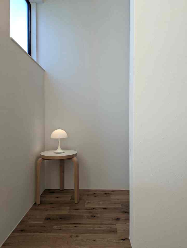 Bright white walls and wood-tone flooring form a cozy corner space with a simple stool topped by a white table lamp, minimalistic interior