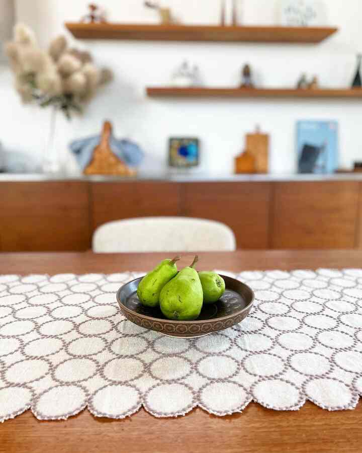 Simple dining space with natural wood tones and white background, featuring a wooden table topped with a circular patterned table runner and green pears