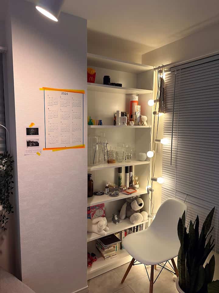 A compact 6畳 room in white tones featuring a poster on the wall, bookshelf, and a white chair next to window blinds with soft lighting