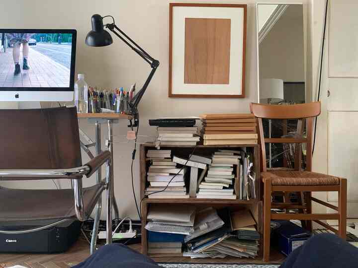 ナチュラルな色調のワークスペース、書籍が積まれた木製ボックスの棚とレザーの椅子、テーブルランプが配置された雰囲気のある空間