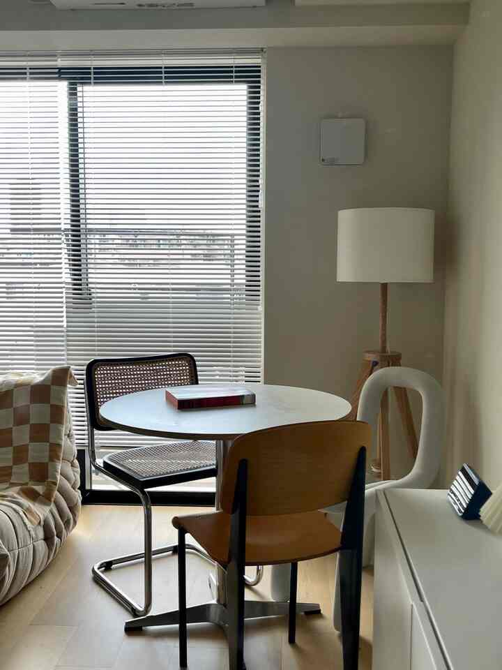 白い壁とブラインドがあるナチュラルな色調のリビング、中央にラウンドテーブルと様々な椅子が配置されたモダンな空間