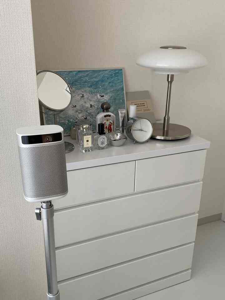 A neat bedroom space in white and silver tones featuring a dresser topped with a table lamp and a projector stand