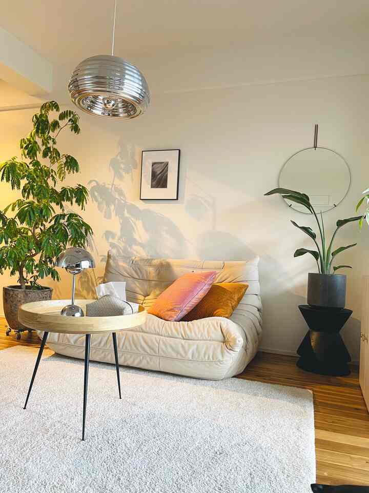 Living room in beige and silver tones featuring a beige sofa center-right, round wooden table with table lamp, and greenery creating a natural atmosphere