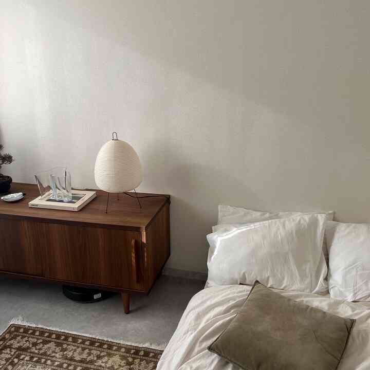 白い壁とナチュラルウッドの家具、シンプルなテーブルランプと寝具が配置されたミニマルなワンルーム寝室の印象的な空間