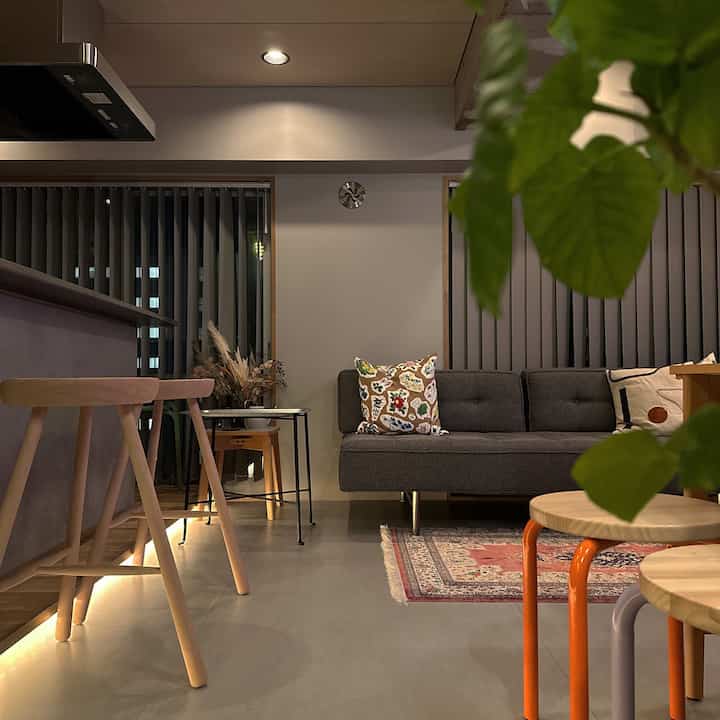 落ち着いたグレーと自然な木目調のリビング、ソファにカラフルなクッション、木製スツールと植物が調和した温かみのあるインテリア