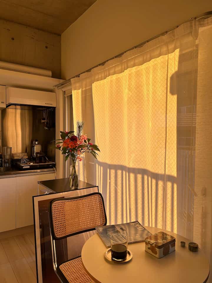 暖かい黄色の光が差し込むワンルームのキッチンとダイニングスペース、UVカーテンと自然素材の家具が配置された雰囲気のあるインテリア