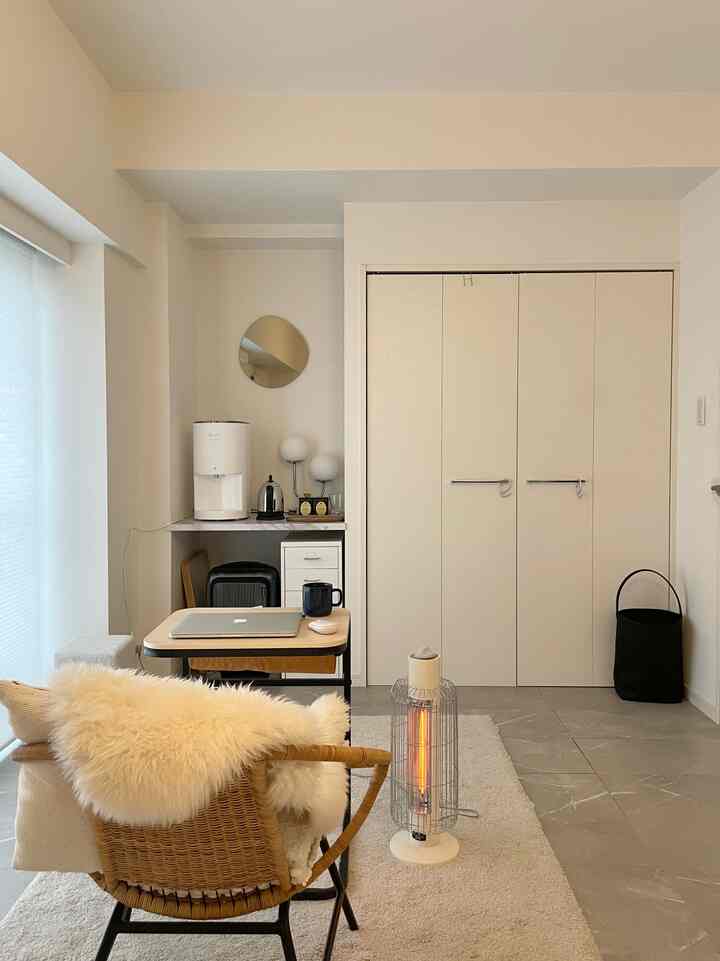 白とベージュのトーンのコンパクトなリビング、ミニマルなデスクとアームチェア、暖房器具が配置された温かみのある空間