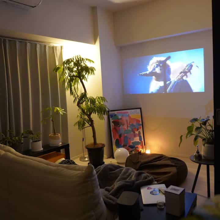 ベージュのソファと複数の観葉植物、ブラウンのビーズクッションがあるナチュラルトーンのリビング、プロジェクター画面でホームシネマの雰囲気を演出