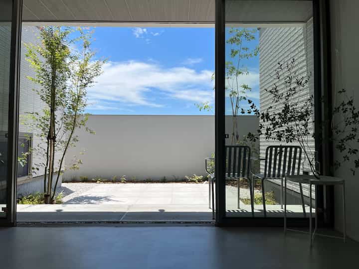 青空と白い壁が織りなす明るいテラス空間、ガラス扉越しに木々とメタルチェアが配されたモダンな室内外の繋がり