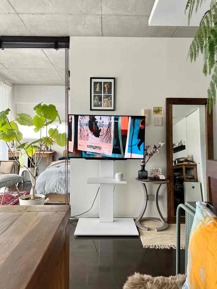白とブラウンのトーンが特徴的な一人暮らしのリビング、中央に電動テレビスタンドとベッドフレーム、緑の植物が置かれた自然な雰囲気のインテリア