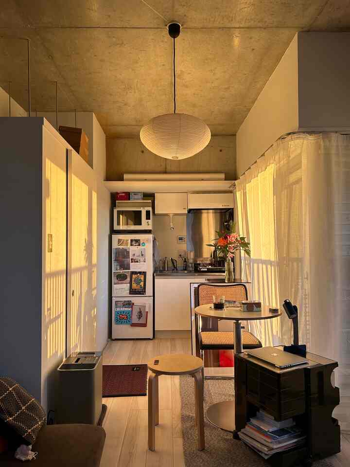 温かい光が差し込むワンルーム、白いキッチンとライトブラウンの床、木製の丸テーブルとスツールが配置された雰囲気のある空間