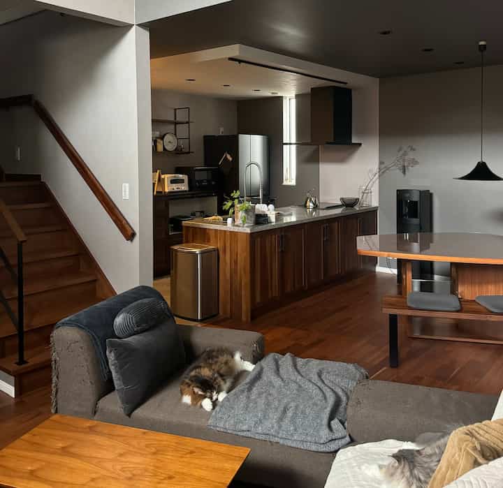 暖かいブラウンの木製とグレーのトーンのリビングとキッチン空間、中央のソファにくつろぐ2匹の猫がいる雰囲気のあるインテリア