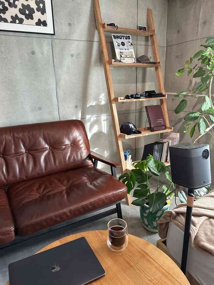 グレーのコンクリート壁とブラウンのレザーソファ、木製棚や植物が配置された落ち着いた一人暮らしのリビング