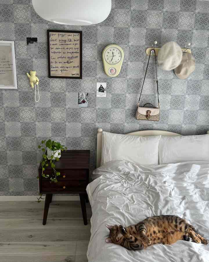 Bedroom with gray floral patterned wall, white bedding, dark wood nightstand, and a cat creating a cozy atmosphere