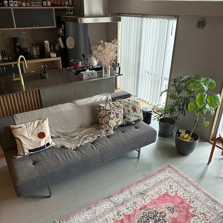 ナチュラルなリビングルーム、グレーのソファと木目調のキッチンアイランド、白い縦型ブラインドが配置された印象的な空間