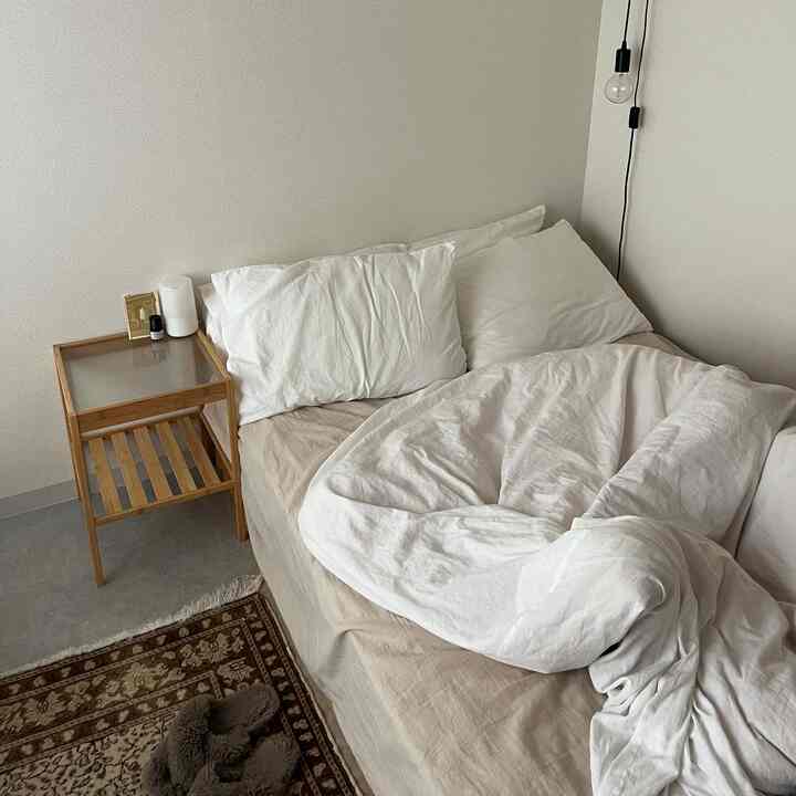 白とベージュのトーンのワンルーム寝室、シンプルな木製ナイトテーブルと整った寝具が配置された落ち着いた空間