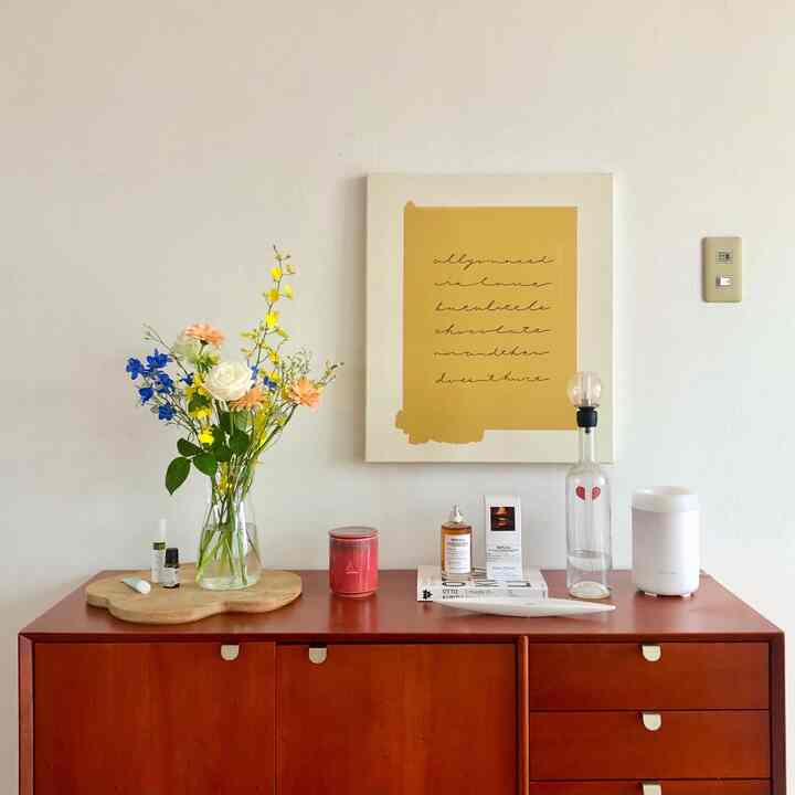 明るいホワイトの壁とナチュラルブラウンの木製キャビネットがあるリビング、花瓶の花とイエロー調のポスターが配置された清潔感のある空間