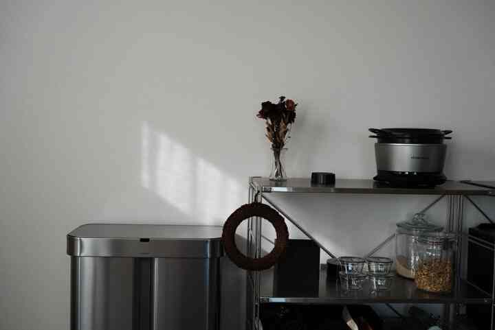 A kitchen space featuring silver metal shelving and trash bin against a white wall illuminated by natural light, creating a clean atmosphere