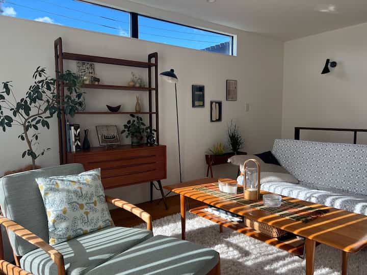 Bright living room with white walls and wood-tone furniture for two, featuring natural light and plants creating a cozy atmosphere