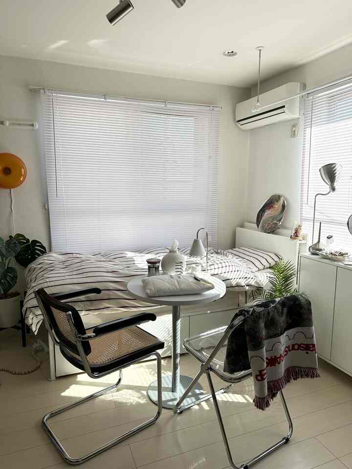 白とナチュラルトーンの小さな寝室、ベッドと丸テーブル、2脚の椅子が配置されたシンプルなミニマルな空間