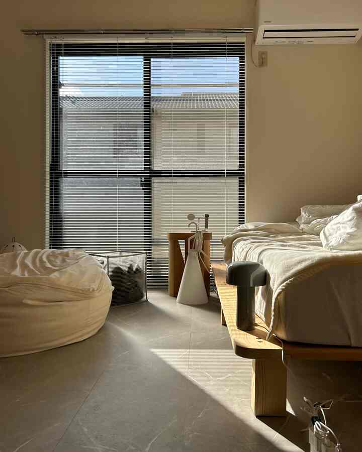 Natural wood tone bedroom featuring large window with blinds, beige bedding, and a white bean bag in a minimalistic space