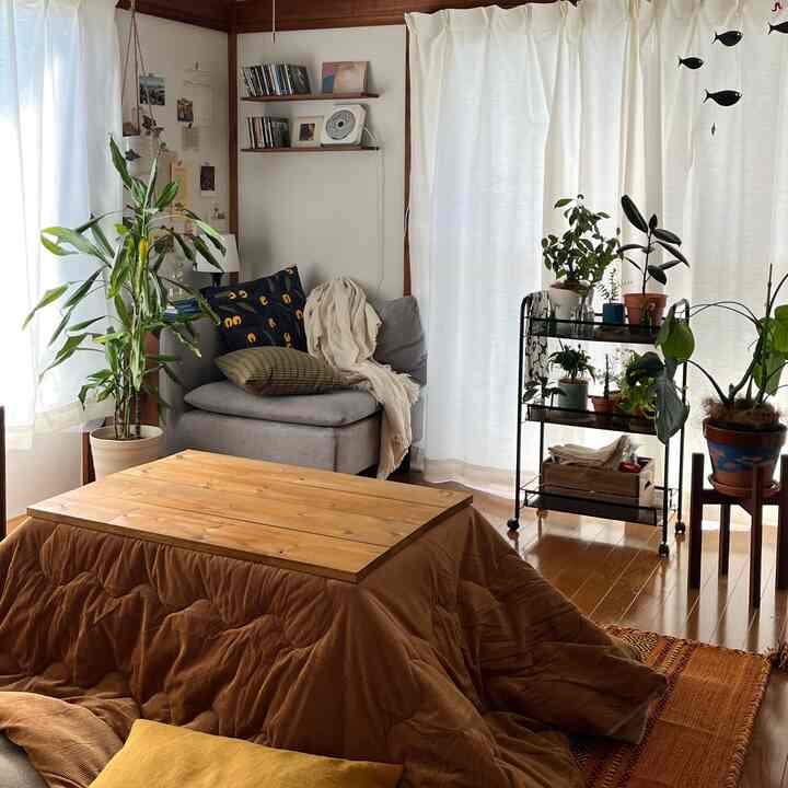 自然なブラウン調のリビング、木製のこたつテーブルとグレーのコーナーソファ、多数の植物が配置された温かみのある空間