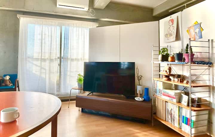 白とナチュラルカラーのワンルームリビング、中央にテレビ台と本棚が配置され、窓にはブラインドがある明るく居心地の良い空間