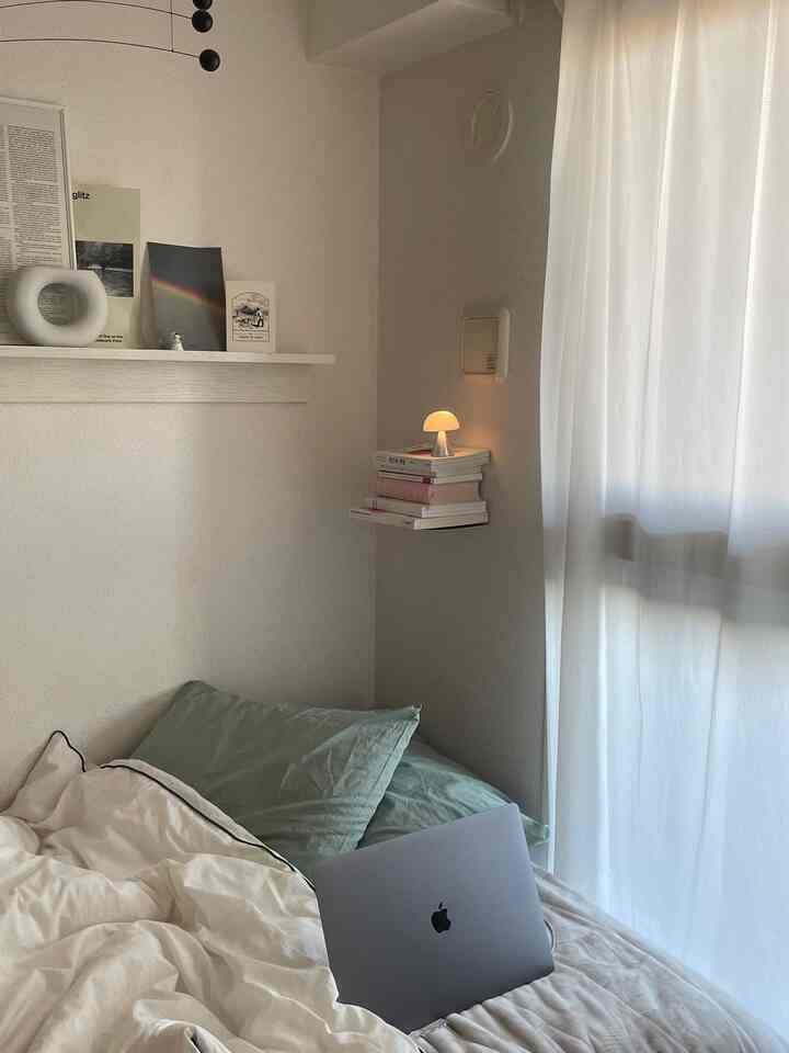 白とナチュラルトーンの寝室、コーナーに設置された本棚と小さなランプが配置された居心地の良いインテリア