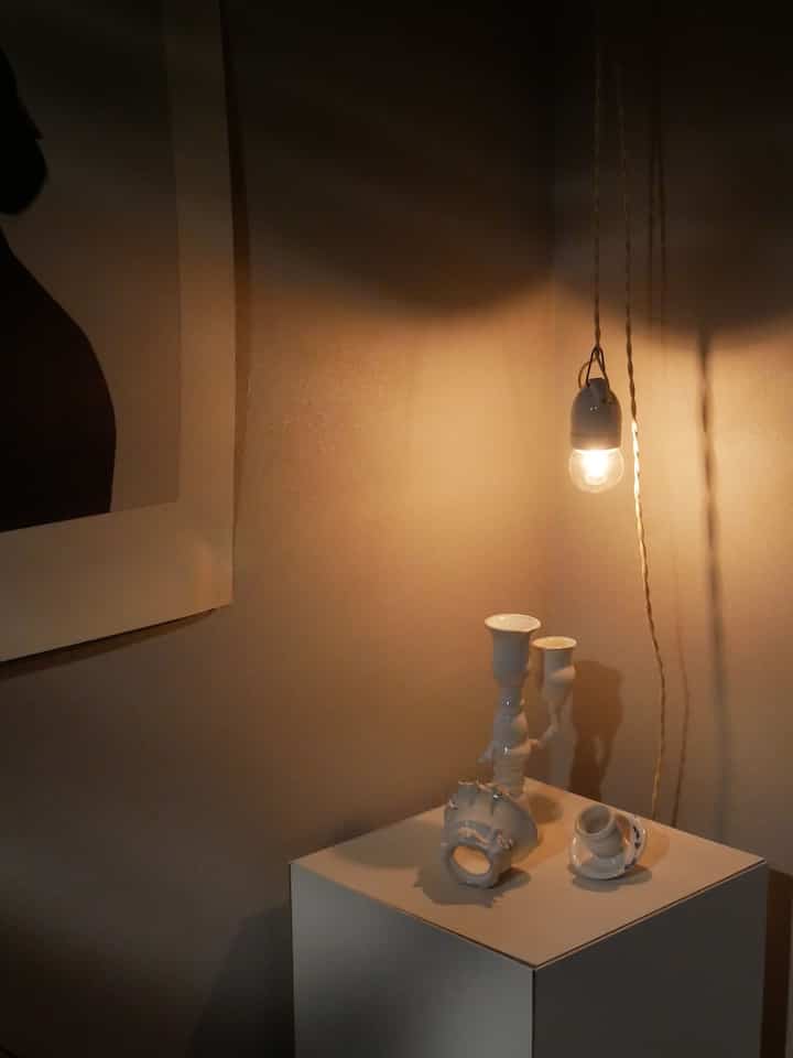 Warm beige-toned entrance corner featuring a display stand with ceramic art objets and a glowing pendant light creating cozy ambiance