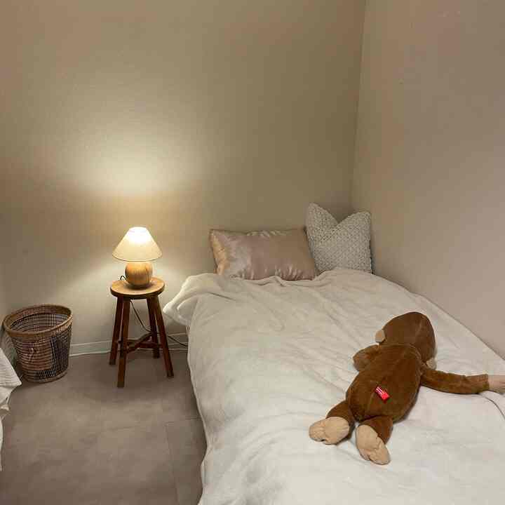 ベージュとブラウンの落ち着いた寝室、クッションとぬいぐるみが置かれたベッドと温かみのあるテーブルランプが配された雰囲気のあるインテリア