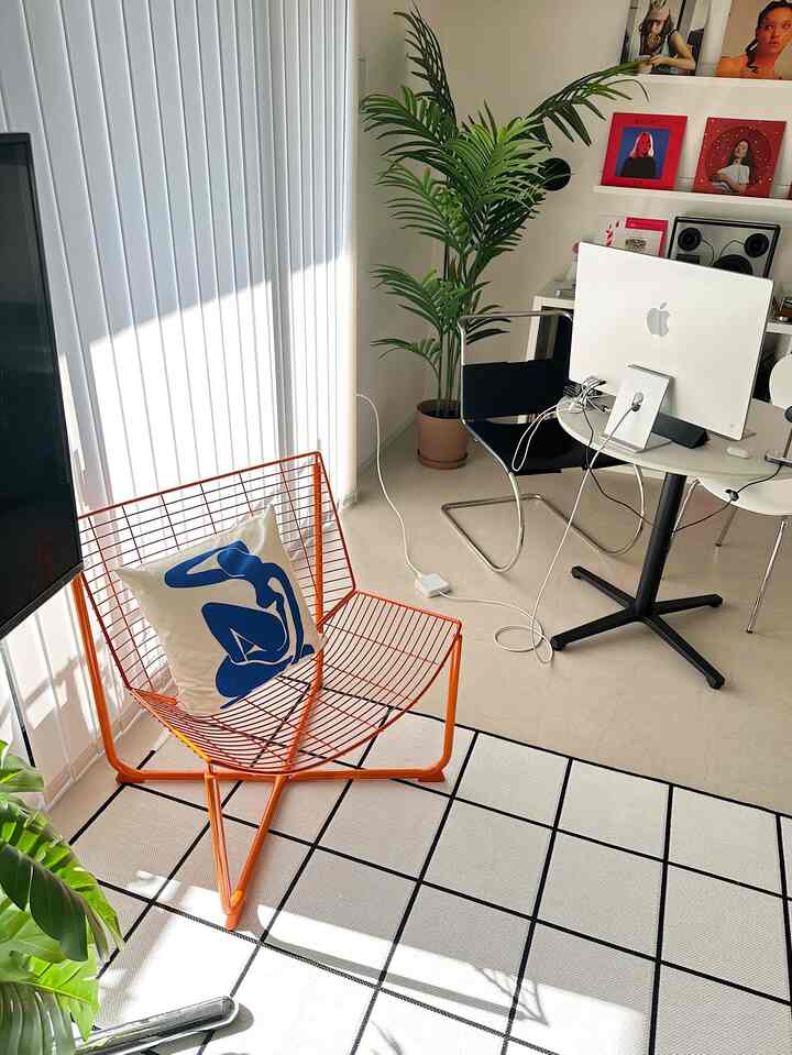 Bright white-toned living room featuring an orange metal armchair, grid-patterned rug, desk with computer, and indoor plants in a mid-century modern style