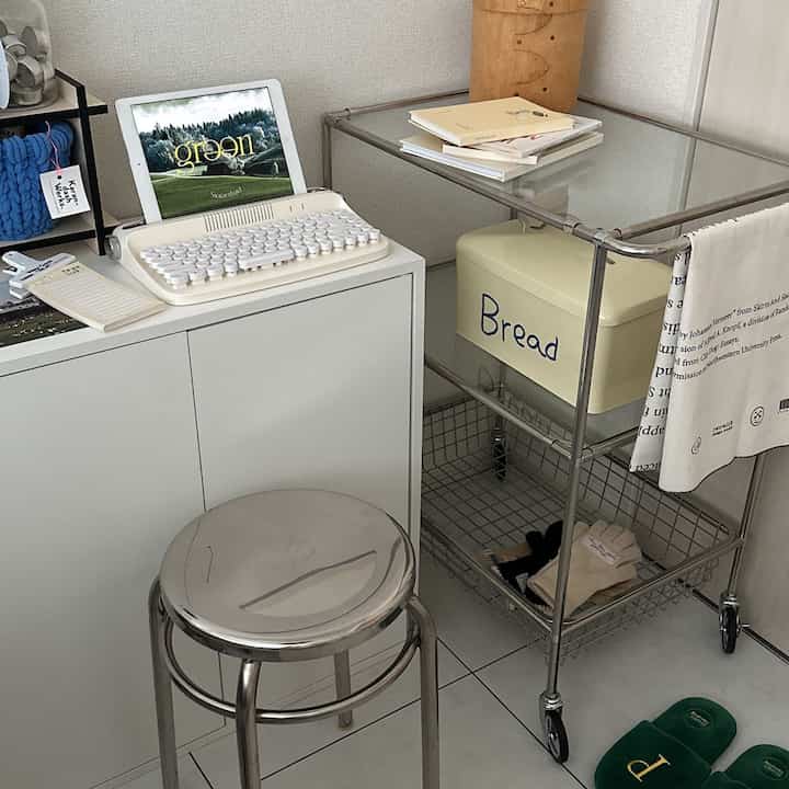White and silver-toned bedroom corner featuring a glass-top trolley and metal stool with a minimal atmosphere