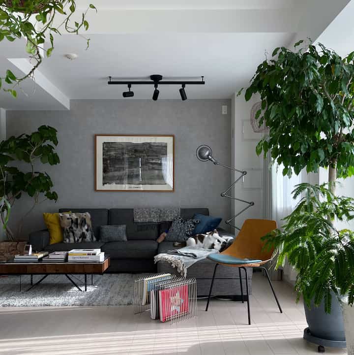 グレーとグリーンのトーンのリビング、広いコーナーソファとラグ、コーヒーテーブル、多数の観葉植物が配置された温かみのある居心地の良い空間